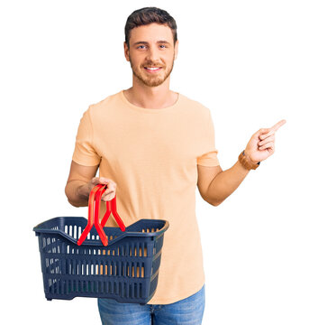 Handsome young man with bear holding supermarket shopping basket smiling happy pointing with hand and finger to the side