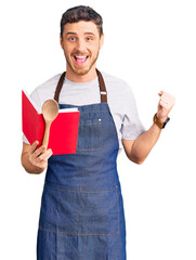 Handsome young man with bear wearing professional baker apron reading cooking recipe book screaming...