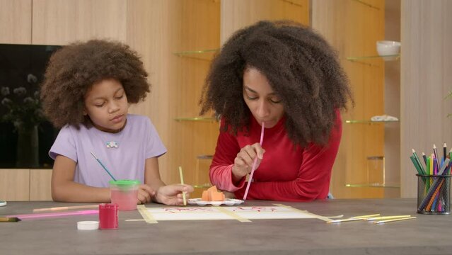 Engrossed adorable elementary age curly black daughter and caring attractive African American mother expressing vivid imagination and fantasy in drawing, blow painting with straws on paper.