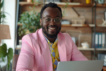 Naklejka premium A man in a pink suit is smiling and sitting in front of a laptop. He is wearing glasses and has a beard. Concept of confidence and professionalism. work portrait of happy black man wearing pink suit