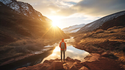 A man stands on a rocky ledge, gazing at the river below as the sun sets. The clouds and sky create...