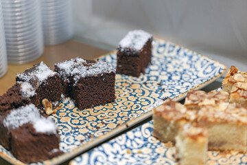Tray of desserts, brownies topped with powdered sugar and almond slices. A cake with almonds is also visible. The tray has a blue and yellow pattern. In the background, there are stacked of cups