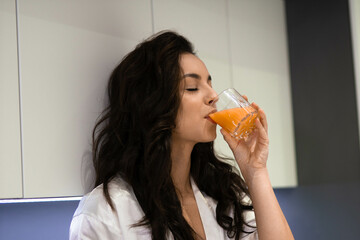 Close up of attractive young Caucasian female with dark long hair in modern kitchen having breakfast holding glass of orange juice. Beautiful young woman drinking fresh juice