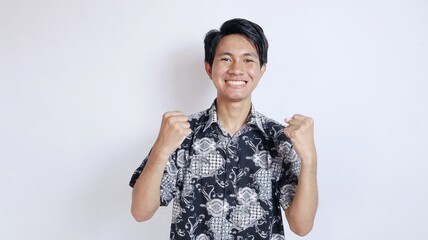 Excited handsome Asian young man wearing batik gesturing celebrating victory and raising