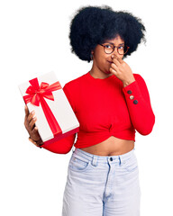 Young african american girl holding gift smelling something stinky and disgusting, intolerable...