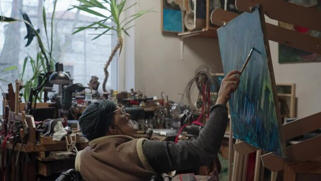Senior artist on a wheelchair paints an oil painting on canvas in a studio