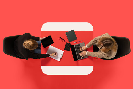 Creative collage. Two young women sit at common table with documents and conduct interview against red studio background. Concept of business, work, job, meeting, office. Copy space. Ad