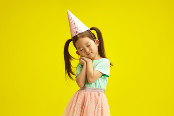 Sweet little girl in party hat dreamly closed eyes, cute placed palms of hands in front of herself...