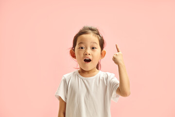 Little Girl with a Surprised Expression Points Up Above her on Free Copy Space, Expresses Joyful and Excited, Showing With Finger Upwards and Looking To Camera with Wide Open Eyes and Mouth