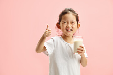 Happy Smiling Little Brunette Child Girl Drinking Milk From Glass With a Trace Frm Milk on Lips, Expresses Pleasure Positively Showing Thumb up Standing On Pink Isolated - 767826183