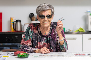 An old woman with wrinkles is sitting in the kitchen while cheerfully smoking a cigarette. - 767828161