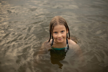 head out of the water, a fair-haired girl looks out of the water after swimming on the lake on a summer evening, relaxation with children on the water, children's sports activity on the water
