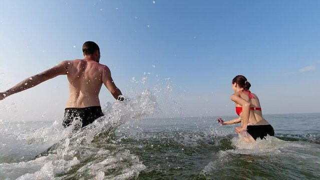 A man and woman run into the sea, kicking up the spray on the first day of their summer vacation. Slow motion. Water games. Vacation and recreation