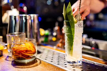 A skilled bartender crafting a refreshing cocktail, adorned with green leaves, next to a whiskey...