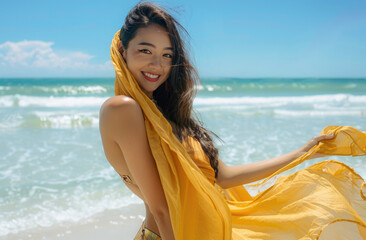 Fototapeta na wymiar A stunning asian woman with long brown hair, wearing an elegant yellow scarf around her waist and shoulder, stands on the beach under clear blue skies