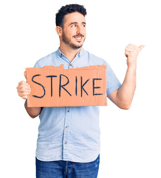 Young hispanic man holding strike banner cardboard pointing thumb up to the side smiling happy with open mouth