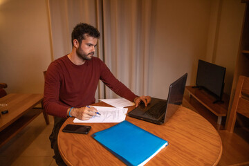 A men working or studing at homen. He is working with a laptop on a desk.