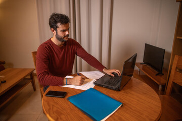 A men working or studing at homen. He is working with a laptop on a desk.