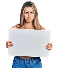 Hispanic young woman holding blank empty banner skeptic and nervous, frowning upset because of...