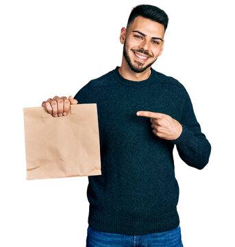 Young hispanic man with beard holding take away paper bag smiling happy pointing with hand and finger