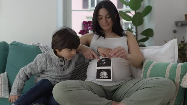 Happy Mother and Child Posing with Ultrasound Picture - Smiling at Camera with Unborn Baby Brother's Image, Seated on Home Couch in Third Trimester