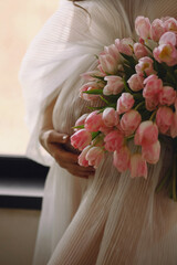 Pregnant woman in a stylish ivory dress holding her belly with one hand a bouquet of pink tulips in...