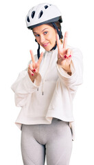 Beautiful brunette young woman wearing bike helmet and sporty clothes smiling looking to the camera showing fingers doing victory sign. number two.