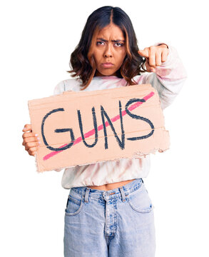 Young beautiful mixed race woman holding no guns warning banner pointing with finger to the camera and to you, confident gesture looking serious