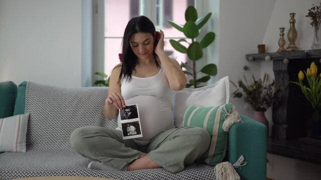 Happy pregnant woman showing ultrasound picture of baby while seated on couch sofa at home during third trimester of pregnancy