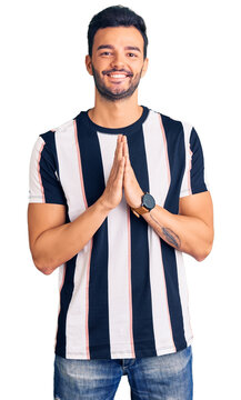 Young handsome hispanic man wearing striped tshirt praying with hands together asking for forgiveness smiling confident.
