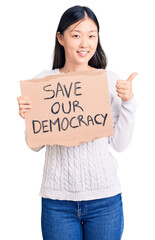 Young beautiful chinese woman holding save our democracy cardboard banner smiling happy and positive, thumb up doing excellent and approval sign