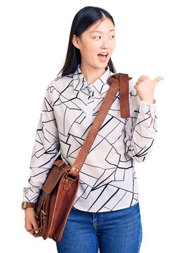 Young beautiful chinese woman holding briefcase pointing thumb up to the side smiling happy with open mouth