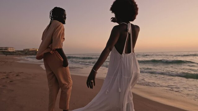 A loving black couple holds hands, walking on the beach at sunset, sharing a moment of joy and romance by the seaside.