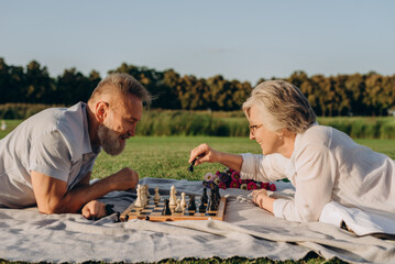 Casually dressed senior couple lying on street rug and competing in game of chess - 767884795