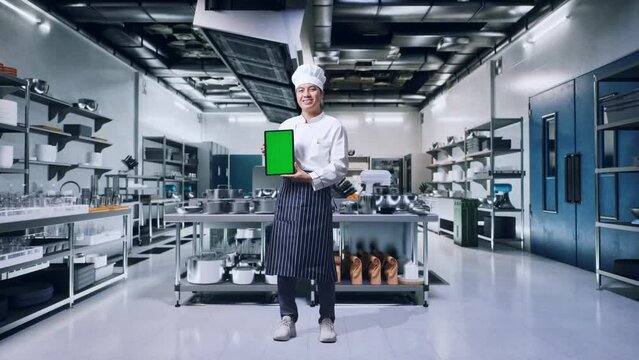 Full Body Of Asian Man Chef Smiling And Showing Green Screen Tablet To The Camera While Standing In Kitchen