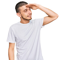Hispanic young man wearing casual white t shirt very happy and smiling looking far away with hand over head. searching concept.