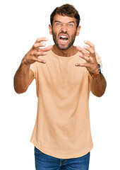 Handsome young man with beard wearing casual tshirt shouting frustrated with rage, hands trying to strangle, yelling mad