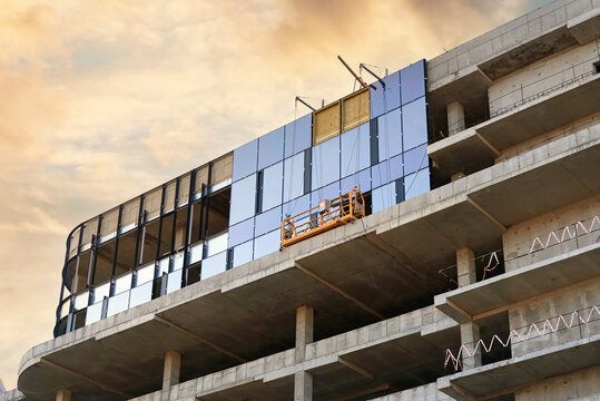 Glazing new construction facade with glass panels. Suspended cradle for facade glazing. Installing glass panes on new building at construction site. Safe glazing processes in urban landscapes