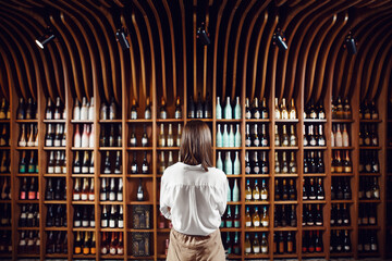 Female sommelier stands in wine cellar with large shelves full of closed bottles with exquisite alcohol drinks put in big collection.