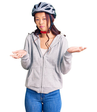 Young beautiful chinese girl wearing bike helmet clueless and confused expression with arms and hands raised. doubt concept.