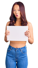 Fototapeta premium Young beautiful chinese girl holding paper banner with blank space scared and amazed with open mouth for surprise, disbelief face