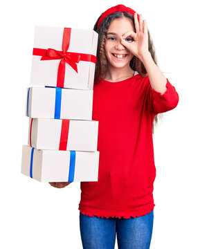 Cute hispanic child girl holding gift smiling happy doing ok sign with hand on eye looking through fingers