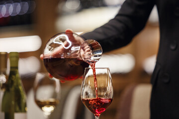 Professional female sommelier pours red wine from decanter to the glass, close up image. Woman waiter pouring alcoholic drink being in cellar of wine shop.