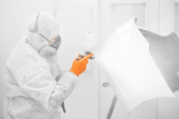Auto mechanic worker painting a white car elements with spray gun in a paint chamber during repair work.