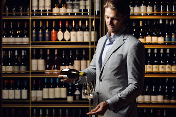 Indoor portrait of male professional sommelier pouring white wine into bottle to the glass being in wine cellar against shelves with various alcoholic beverages. Wine school, winemaking master class.