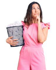 Young beautiful brunette woman holding paper bin full of crumpled papers serious face thinking about question with hand on chin, thoughtful about confusing idea