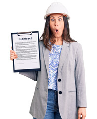 Young beautiful brunette woman wearing architect hardhat holding contract scared and amazed with open mouth for surprise, disbelief face