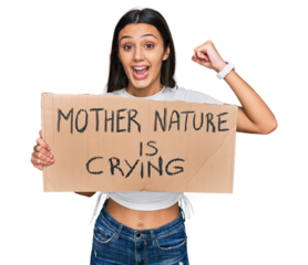 Young hispanic girl holding mother nature is crying protest cardboard banner screaming proud, celebrating victory and success very excited with raised arms