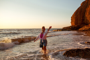 A beautiful plus size woman dressed solid  pink swimsuit, jeans shorts, sunglasses and white shirt walks along the seashore against the sand beach and sea or ocean during the sunset