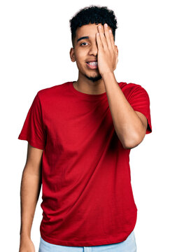 Young african american man wearing casual red t shirt covering one eye with hand, confident smile on face and surprise emotion.